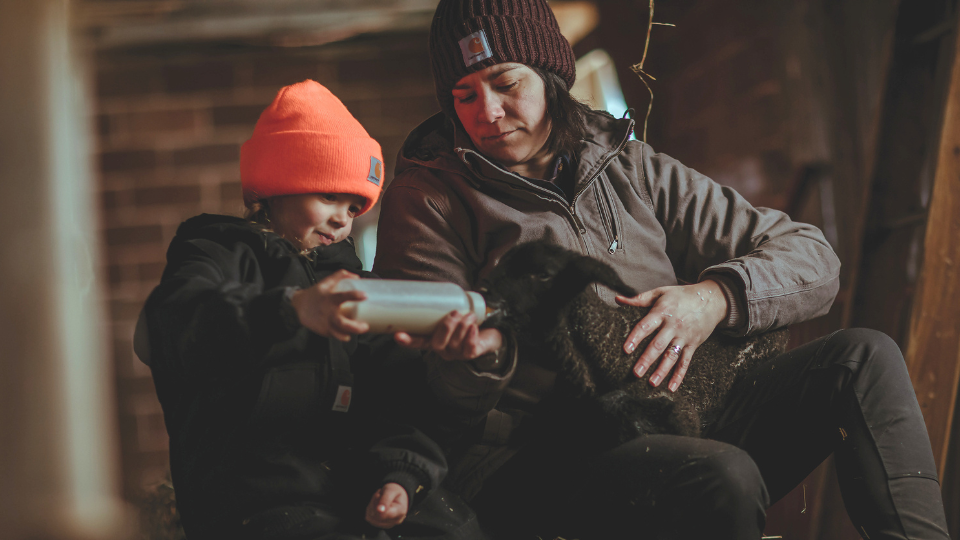A mother and daughter feeding a baby lamb while wearing Carhartt clothing.